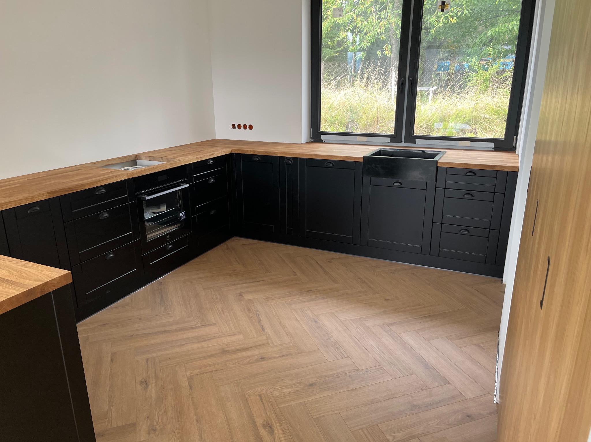 Nowoczesna kuchnia z czarnymi szafkami i drewnianym blatem, widok na okno z zielenią. Parkiet w jodełkę dodaje elegancji. Minimalistyczny design.