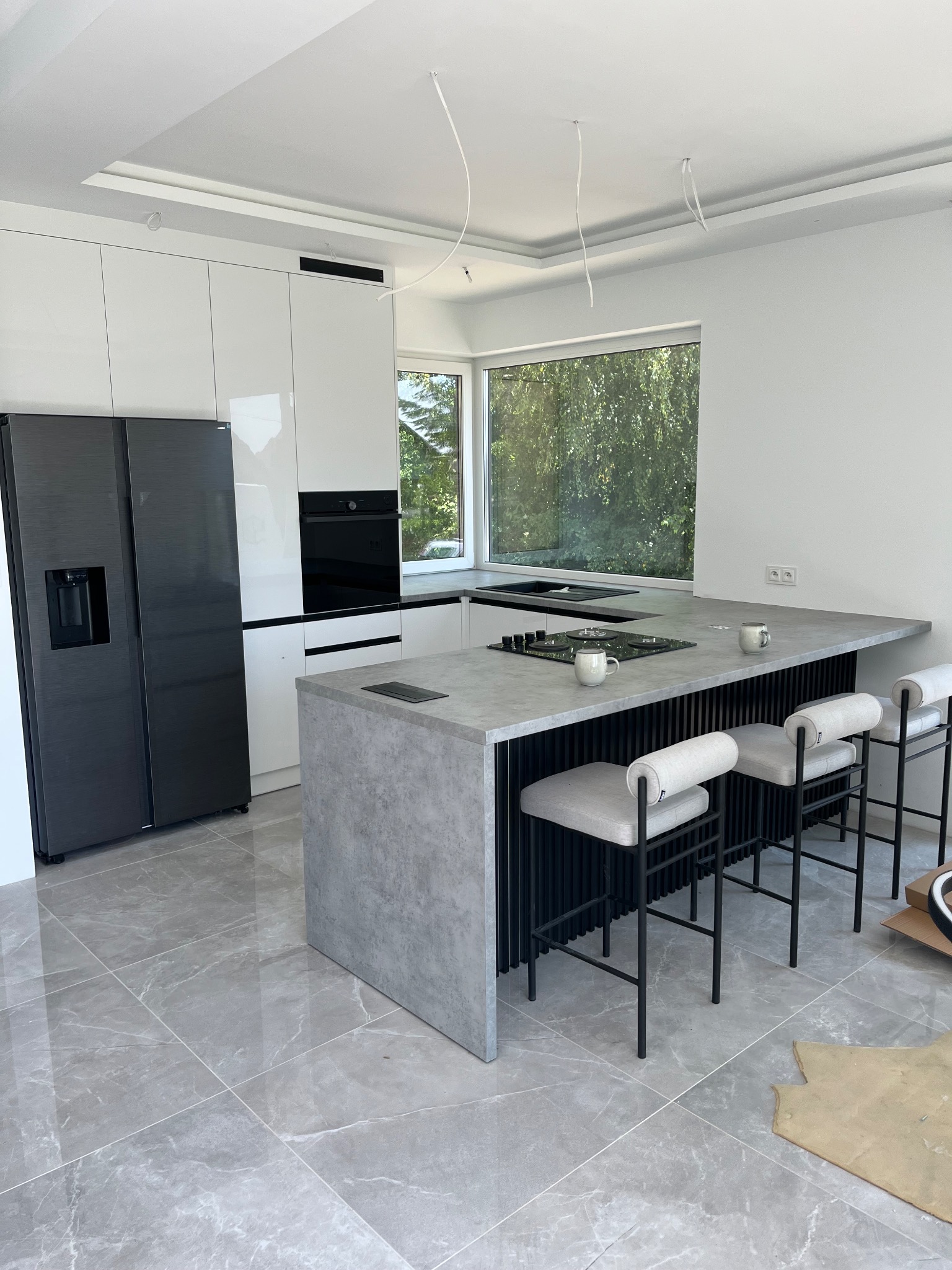Nowoczesna kuchnia z wyspą i hokerami, wykończona w bieli i szarości. Widoczny czarny sprzęt AGD i marmurowe płytki podłogowe. Minimalistyczny design z elementami dekoracyjnymi.