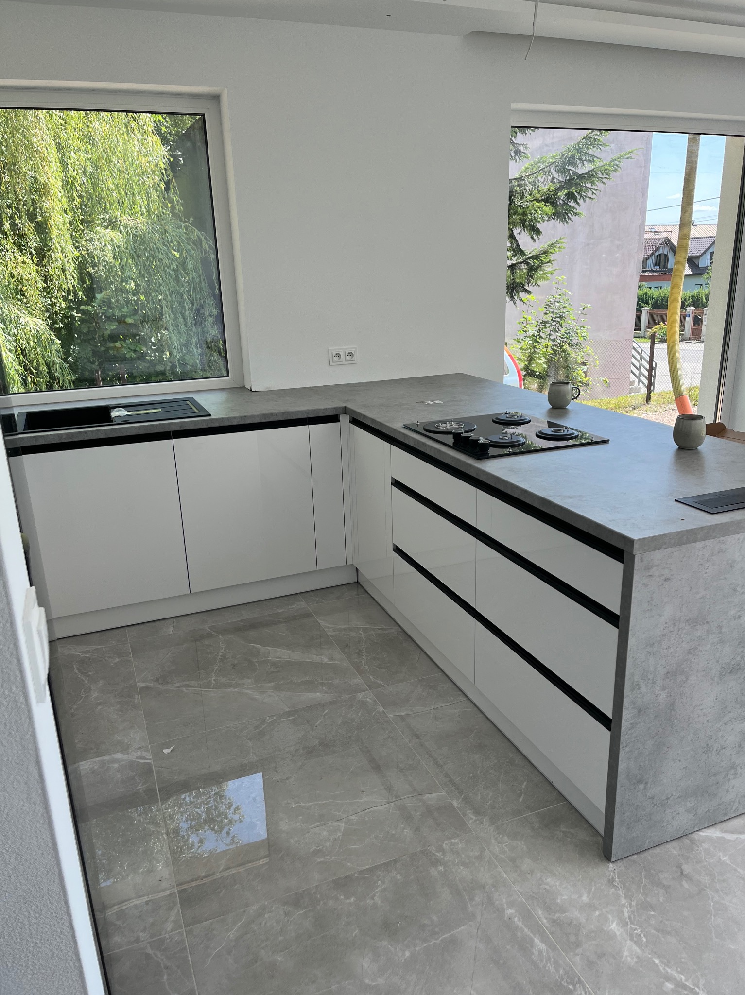 Biała kuchnia z szarym blatem i płytą grzewczą, widok na zieleń za oknem. Minimalistyczny design, marmurowe płytki podłogowe. Nowoczesne wykończenie.