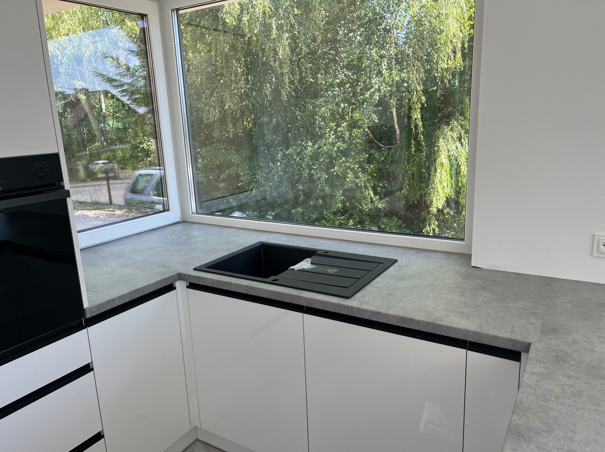 Nowoczesna kuchnia z białymi szafkami i szarym blatem, czarny zlew pod oknem z widokiem na zieleń. Minimalistyczny design, jasne wnętrze, świeżo po montażu.