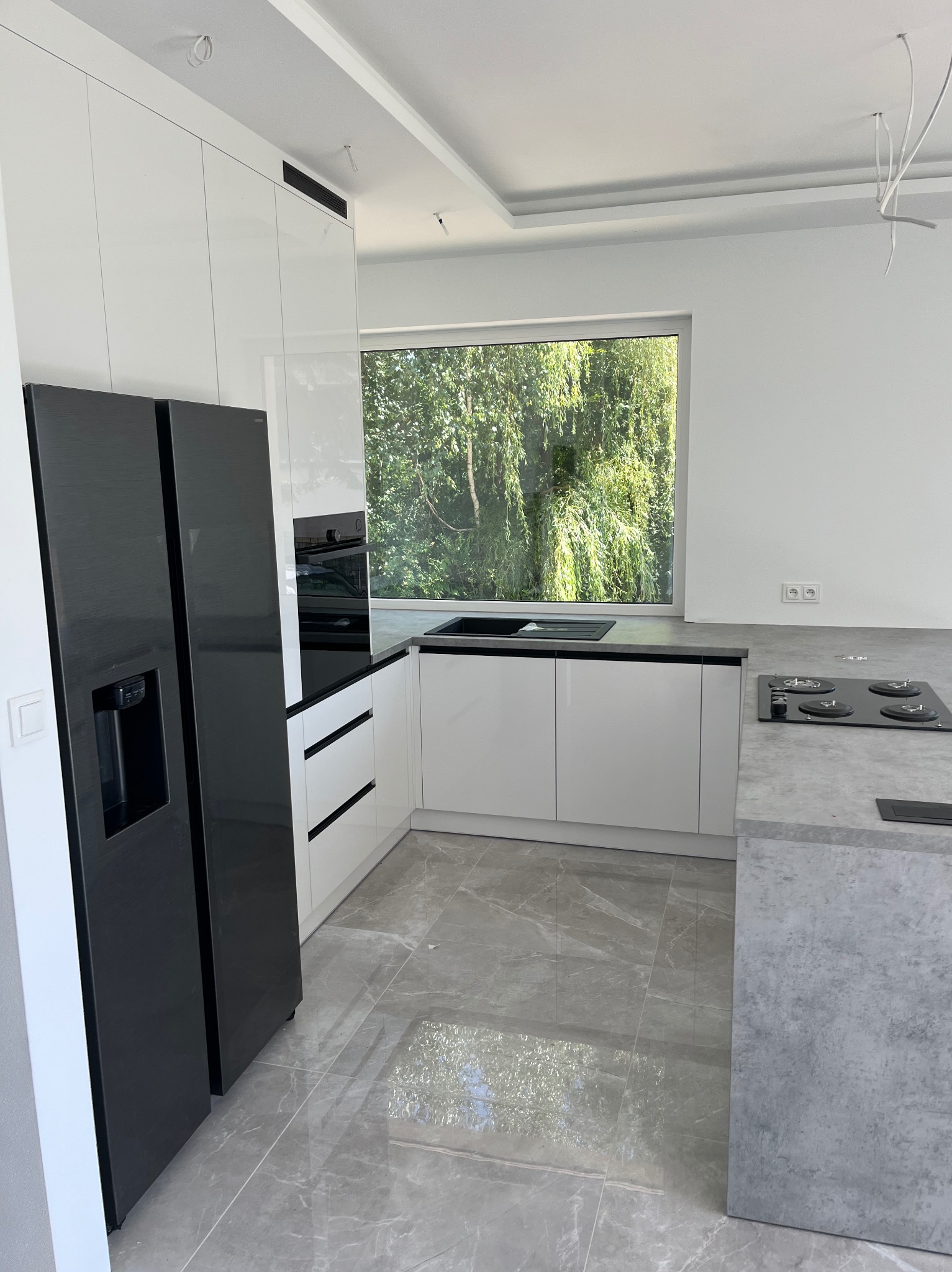 Nowoczesna, jasna kuchnia z białymi szafkami i czarnym sprzętem AGD. Duże okno z widokiem na zieleń. Szare płytki podłogowe z marmurkowym wzorem.