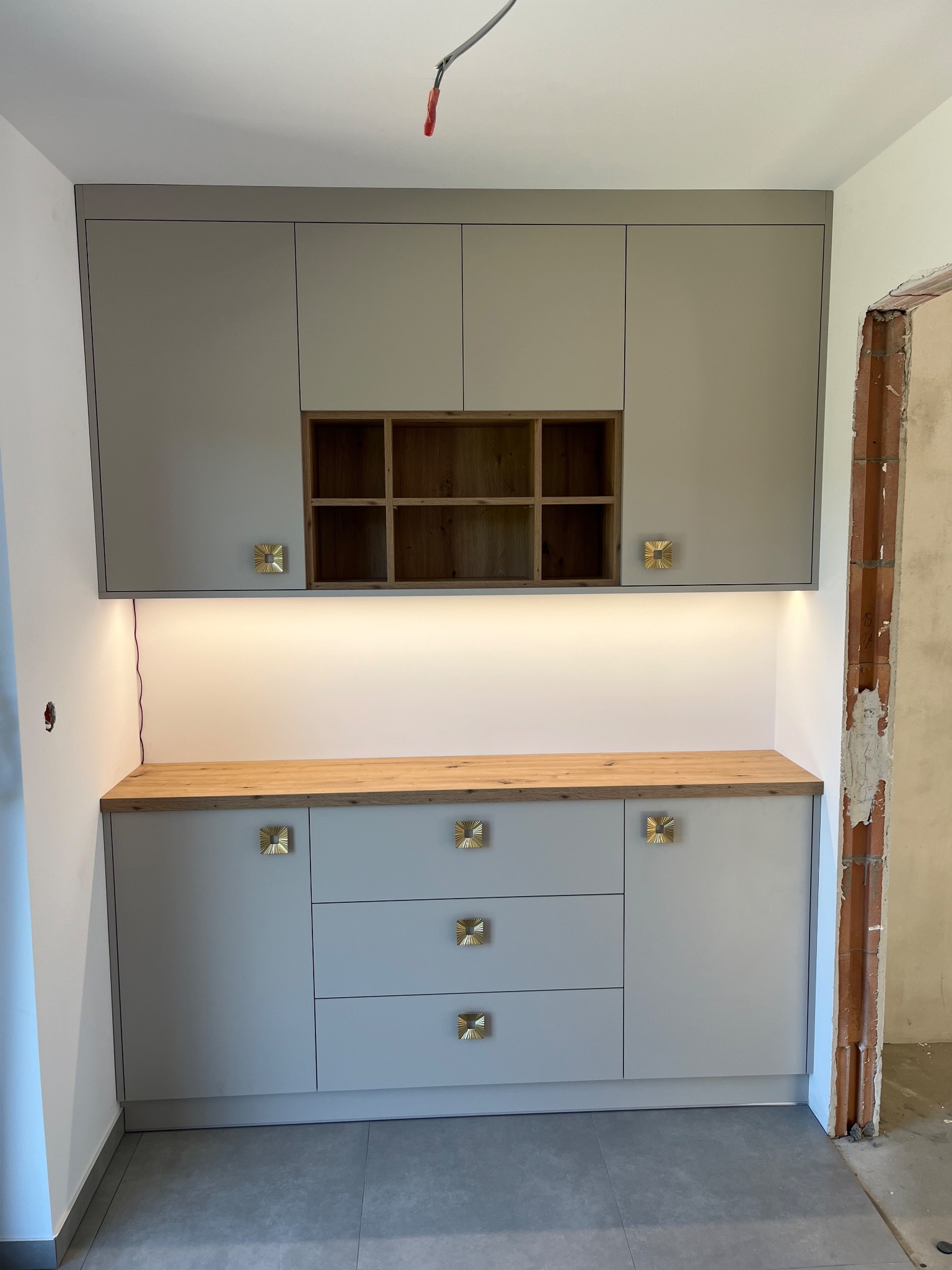 Jasnoszara zabudowa kuchenna z drewnianym blatem i złotymi uchwytami, oświetlona ledami, na tle surowego muru. Widoczne trzy szuflady i szafki wiszące.