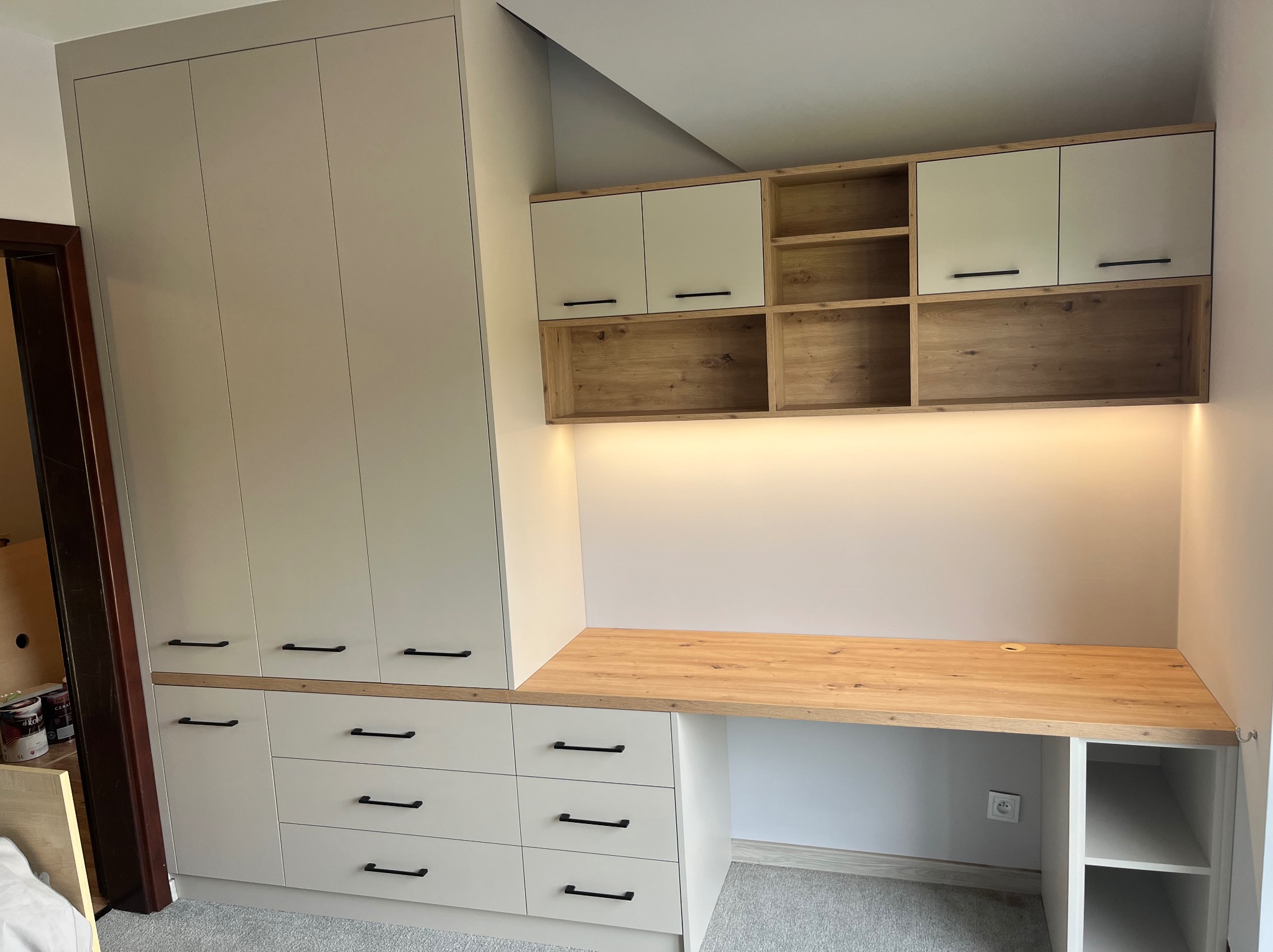 Jasna zabudowa meblowa z szafą, szafkami wiszącymi i biurkiem z blatem drewnopodobnym, czarne uchwyty, oświetlenie LED pod szafkami wiszącymi.