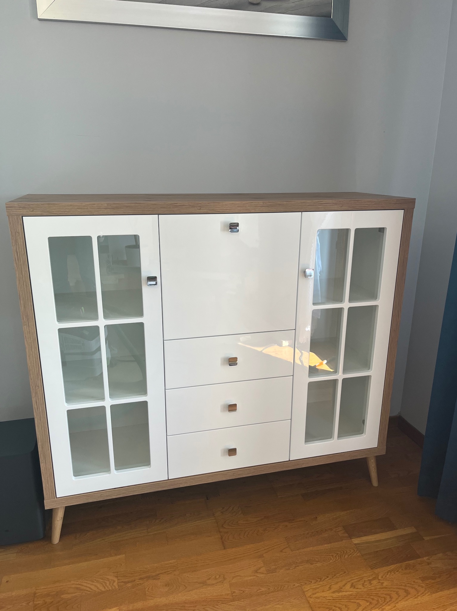 Biało-drewniana komoda z przeszklonymi drzwiczkami i trzema szufladami, styl retro, na drewnianej podłodze. Widoczne detale wykończenia i uchwyty.