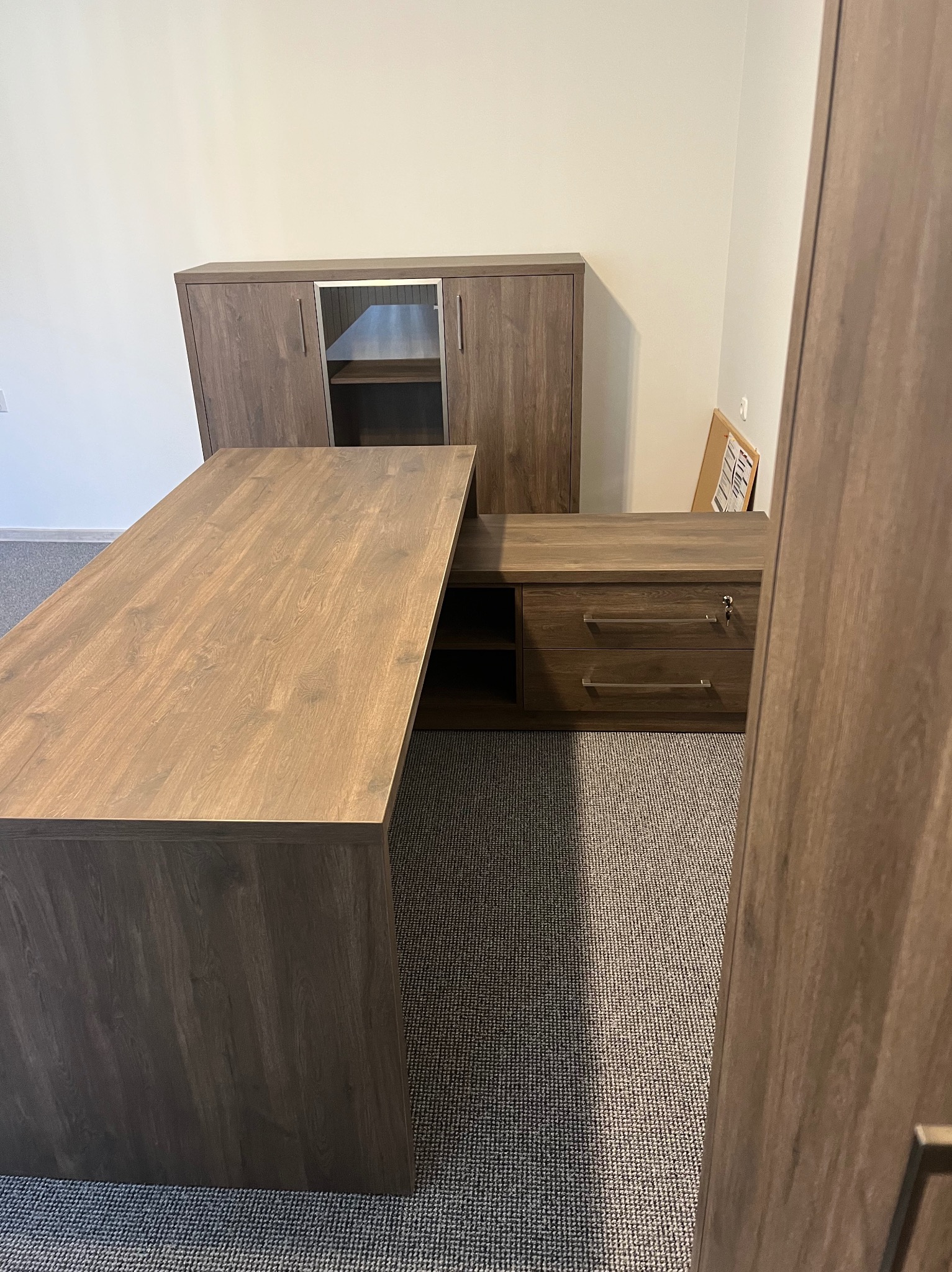 Kątowe biurko z szafkami w odcieniach brązu, z widoczną strukturą drewna, na tle szarej wykładziny. Elementy biurowe w tle.