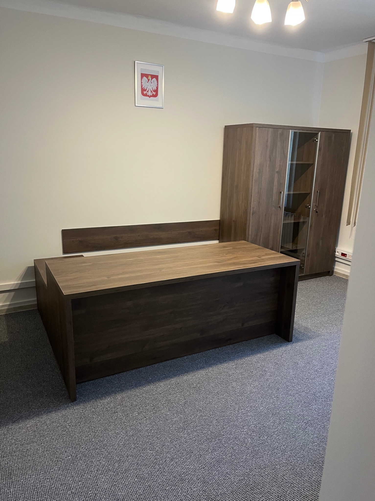 Biuro z ciemnobrązowym biurkiem i szafą z przeszkleniami, na ścianie godło Polski. Wykładzina dywanowa w szarym odcieniu. Oświetlenie sufitowe.