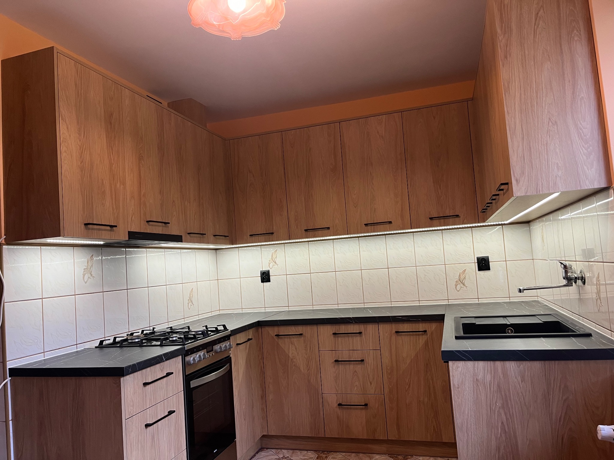 Jasna kuchnia z drewnianymi frontami, czarnym blatem i płytkami z motywem kwiatowym. Oświetlenie LED podszafkowe. Ujęcie z narożnika, widoczna kuchenka i zlew.