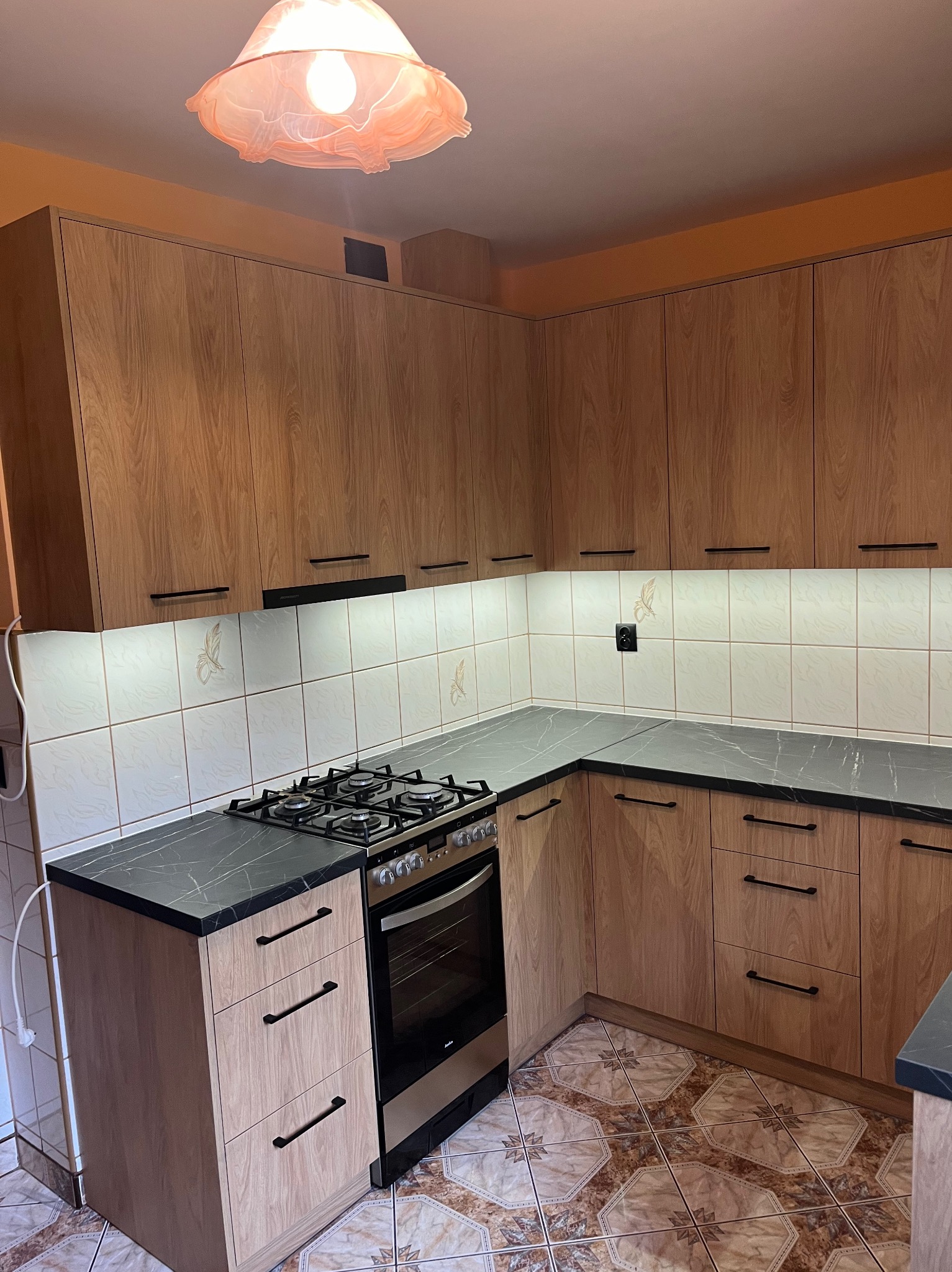 Jasne drewniane szafki kuchenne z czarnymi uchwytami i blatem, oświetlenie podszafkowe, wzorzyste płytki podłogowe, kuchenka gazowa z piekarnikiem.