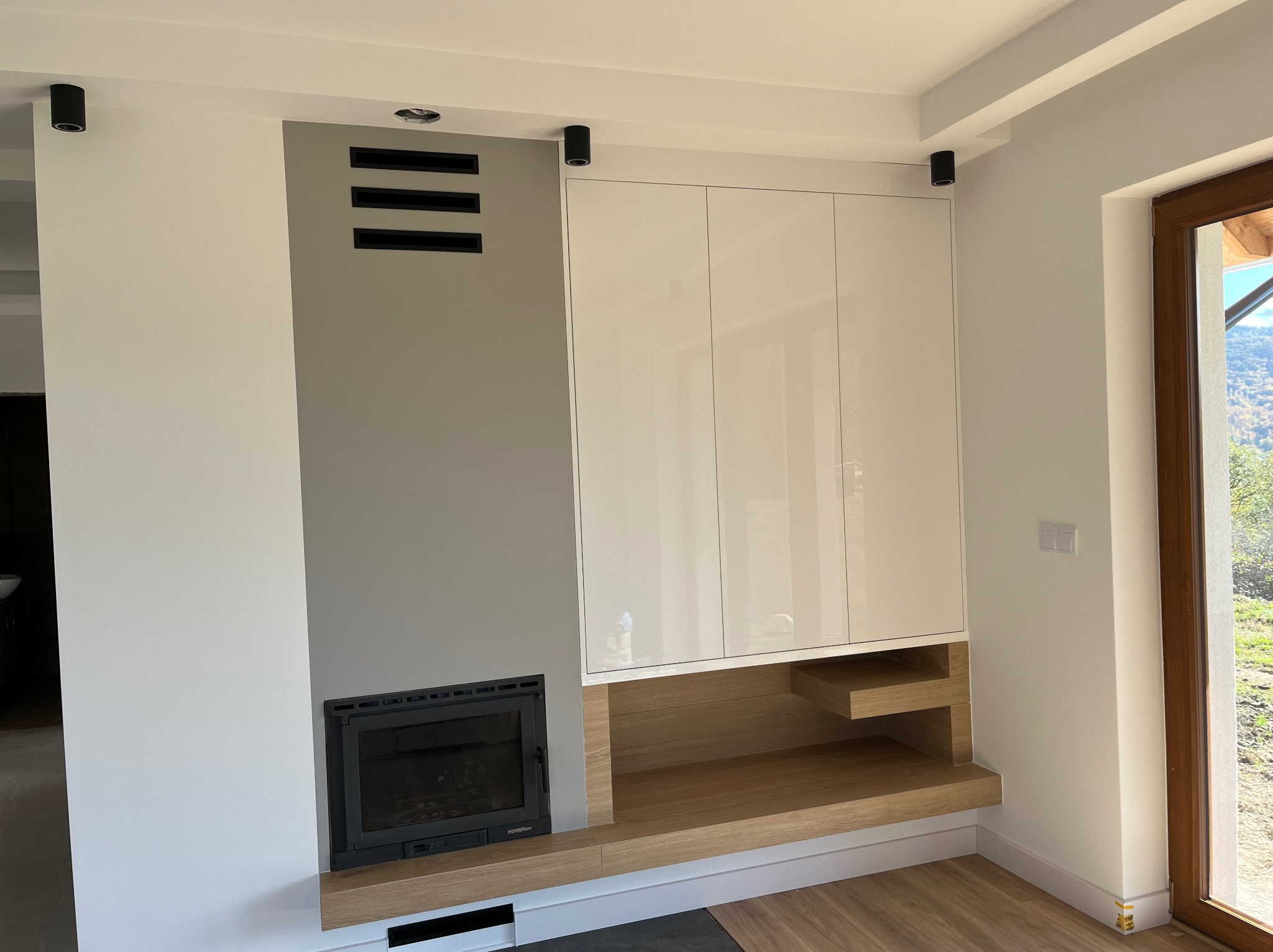 Biała szafa wnękowa z drewnianymi półkami, wkomponowana obok kominka w szarej wnęce. Minimalistyczny design, oświetlenie punktowe.
