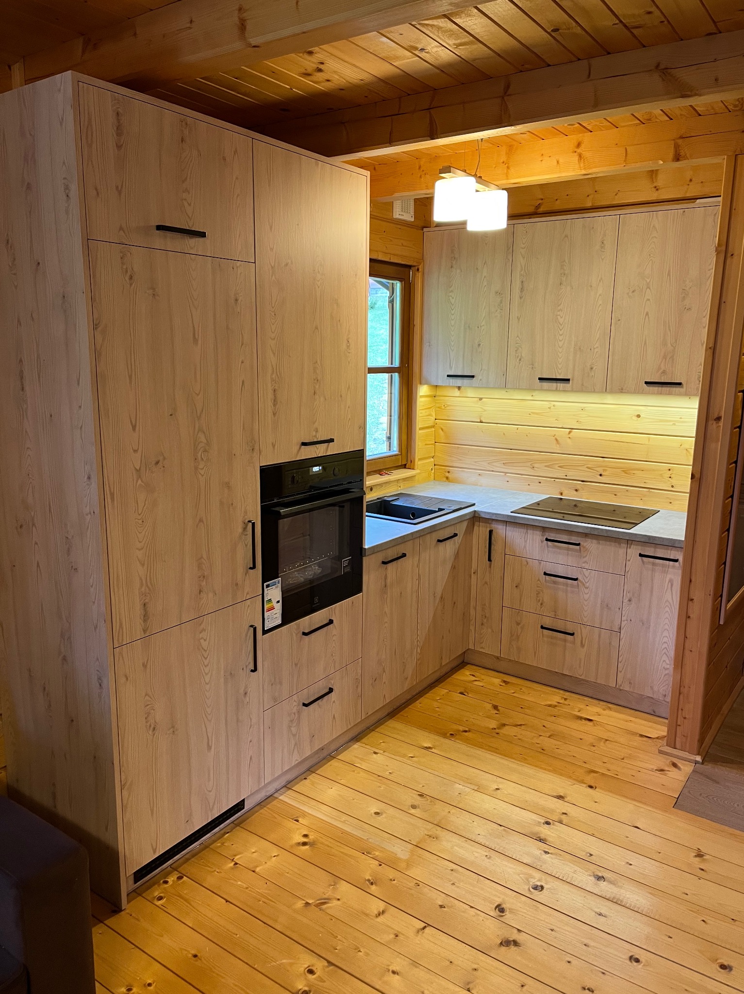 Drewniana kuchnia w rustykalnym stylu z czarnymi uchwytami, wbudowanym piekarnikiem i oświetleniem LED pod szafkami, w drewnianym domu.