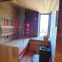 Wnętrze drewnianej kabiny z przeszklonymi drzwiami, podświetlane ławki i panele grzewcze na ścianach, piec z kamieniami i drewniane wiadro z chochlą. Widok na zewnątrz przez otwarte drzwi.