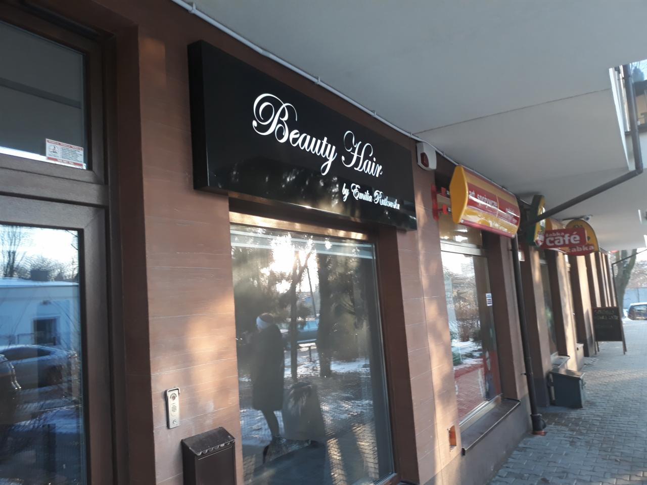 Czarny szyld reklamowy z białym napisem 'Beauty Hair by Emilia Rutkowska' nad wejściem do salonu, widoczny z zewnątrz na tle chodnika i innych lokali.