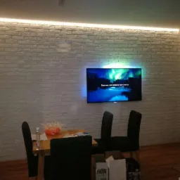 Pokój dzienny z białymi cegłami na ścianie, podświetlonym sufitem LED i wiszącym telewizorem wyświetlającym zorzę polarną, nad stołem z krzesłami.
