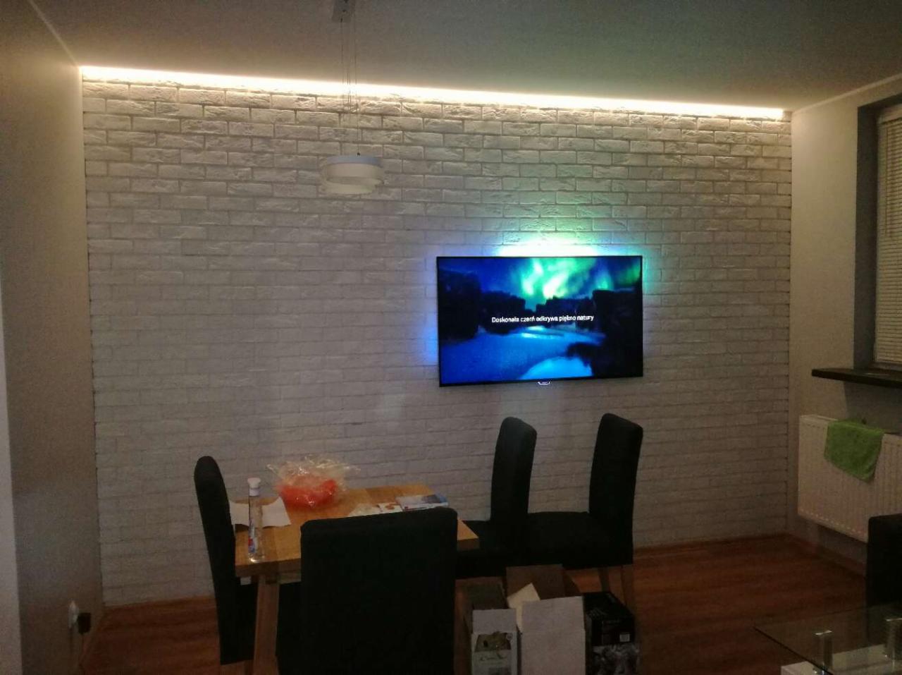 Pokój dzienny z białymi cegłami na ścianie, podświetlonym sufitem LED i wiszącym telewizorem wyświetlającym zorzę polarną, nad stołem z krzesłami.