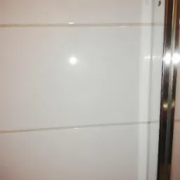 Białe, błyszczące płytki na ścianie kabiny prysznicowej z widocznymi fugami i metalową ramą drzwi.