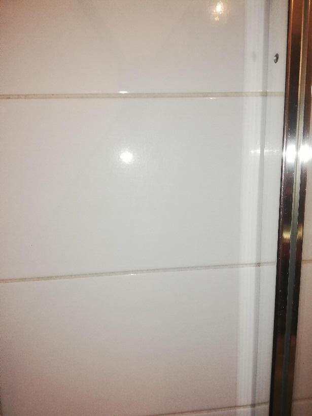 Białe, błyszczące płytki na ścianie kabiny prysznicowej z widocznymi fugami i metalową ramą drzwi.