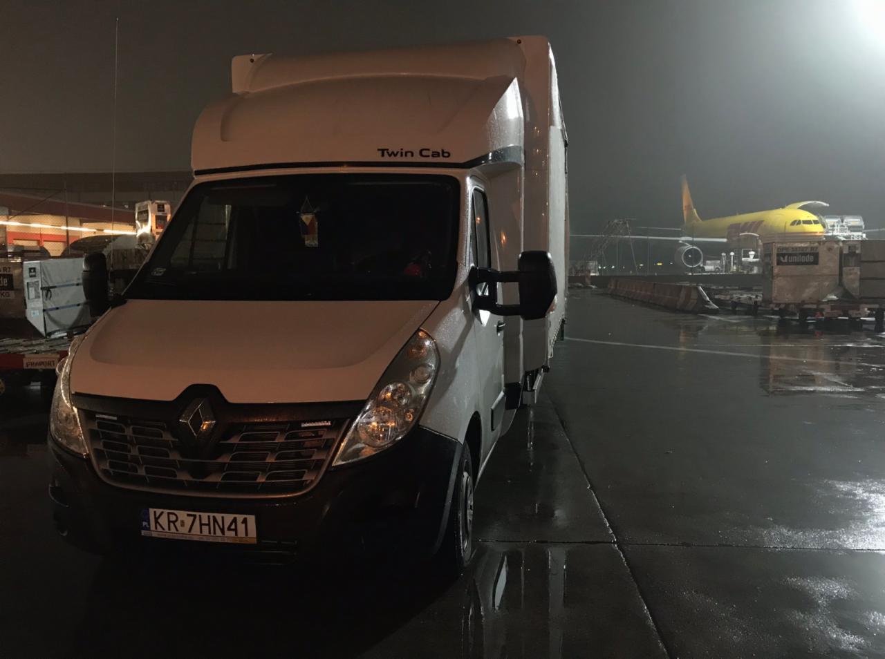 Biały bus Renault Twin Cab na mokrym asfalcie lotniska, w tle żółty samolot transportowy DHL i lampy oświetlające płytę