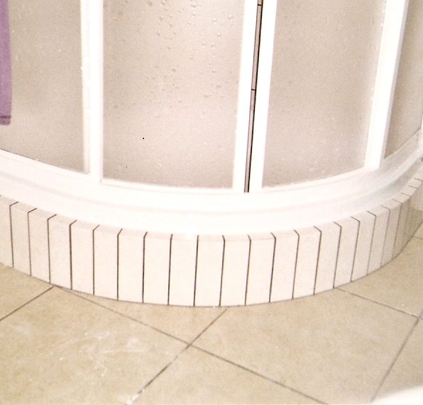 Półokrągła kabina prysznicowa z matowego szkła i białymi ramami, wykończona rzędem pionowych płytek na podstawie, widoczna na tle płytek podłogowych.