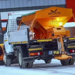 Pojazd specjalny z pomarańczowym osprzętem do usuwania śniegu, widok z tyłu, na śnieżnym podłożu, z widocznym oświetleniem ostrzegawczym.