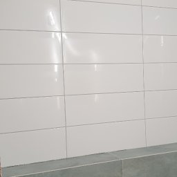 Ps-bau - Ściana łazienki z białymi, błyszczącymi płytkami ceramicznymi ułożonymi poziomo, z widocznym otworem wentylacyjnym w górnym rogu oraz fragmentem dolnej części ściany wykończonej w szaro-zielonych...