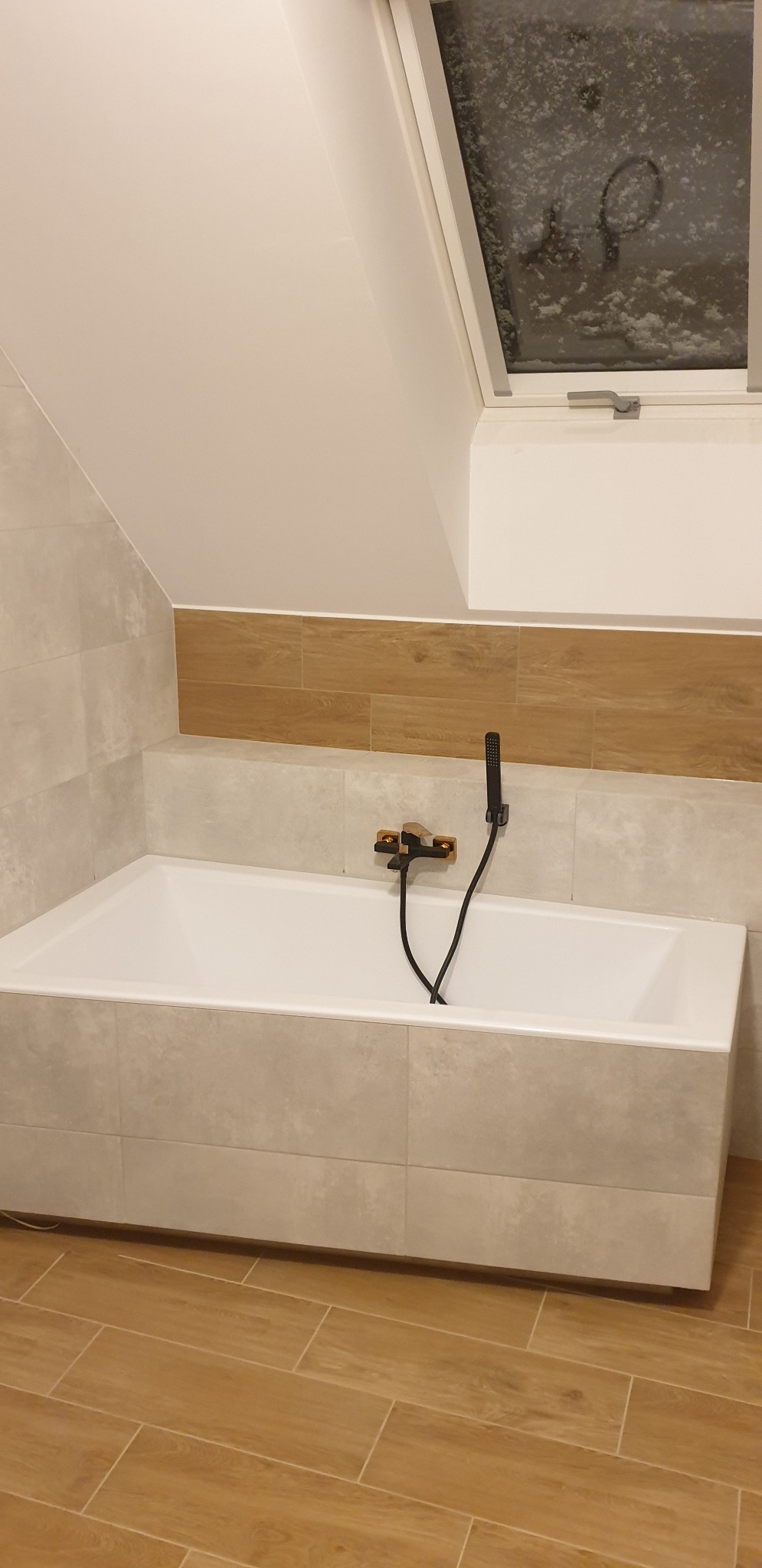 Nowoczesna łazienka na poddaszu z białą wanną obłożoną płytkami, drewnopodobnymi płytkami na ścianie i podłodze oraz czarną armaturą, widok na okno dachowe z zewnątrz pokryte śniegiem.