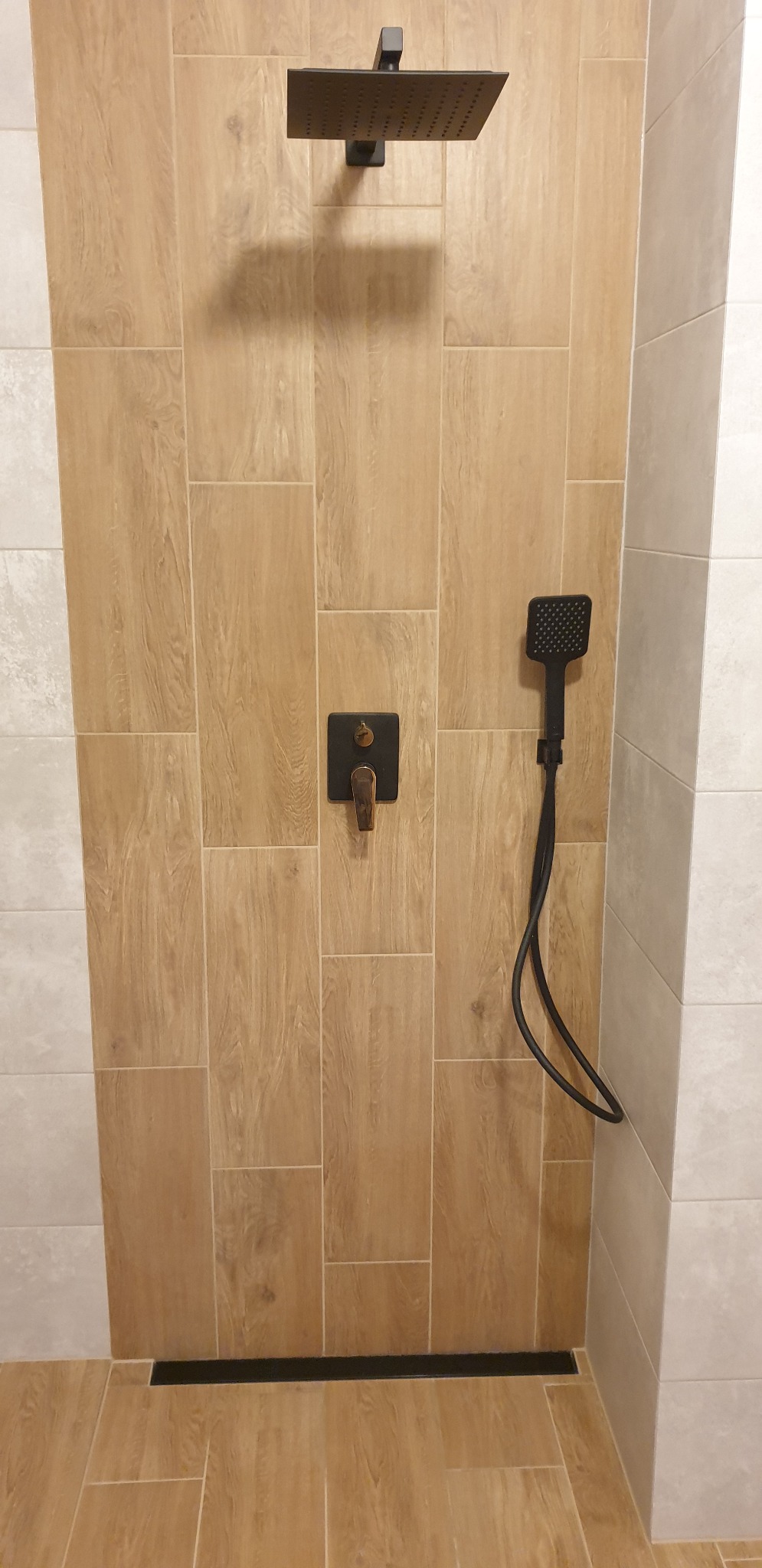 Nowoczesna kabina prysznicowa z deszczownicą w kolorze czarnym, wykończona płytkami imitującymi drewno i szarymi płytkami, z odpływem liniowym.