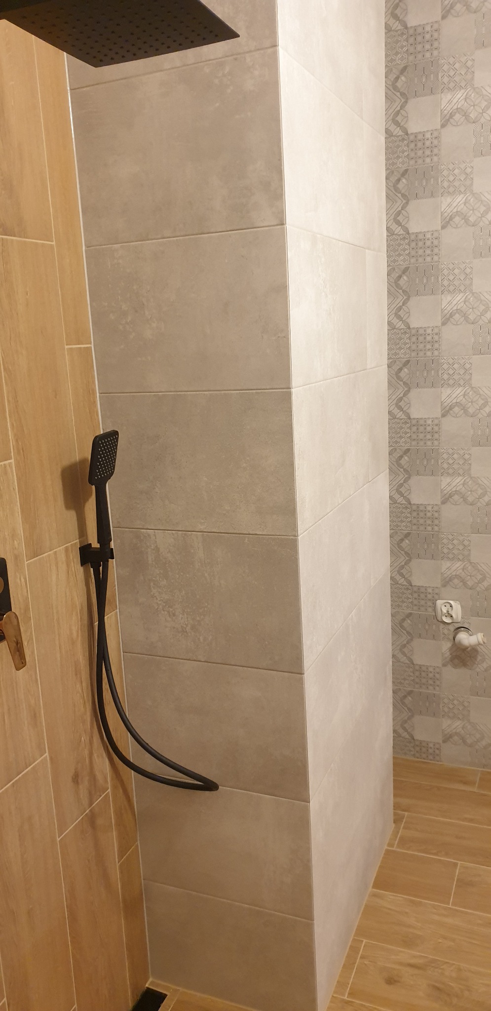 Pionowy kadr na kabinę prysznicową z deszczownicą i słuchawką w kolorze czarnym, wykończoną płytkami w odcieniach beżu i szarości z delikatnym wzorem na jednej ze ścian.