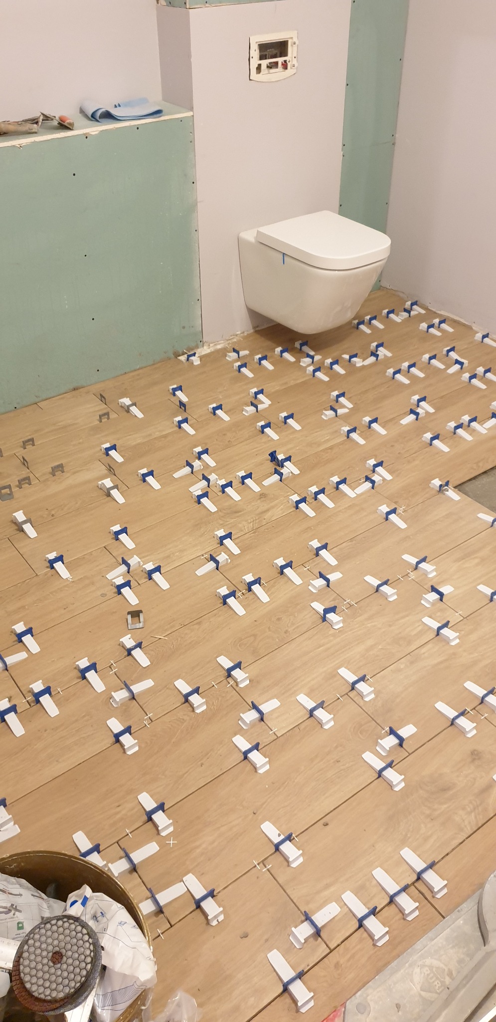 Układanie płytek imitujących drewno w łazience, widoczne białe klipsy poziomujące z niebieskimi klinami, wisząca toaleta w tle, ściany przygotowane pod malowanie.