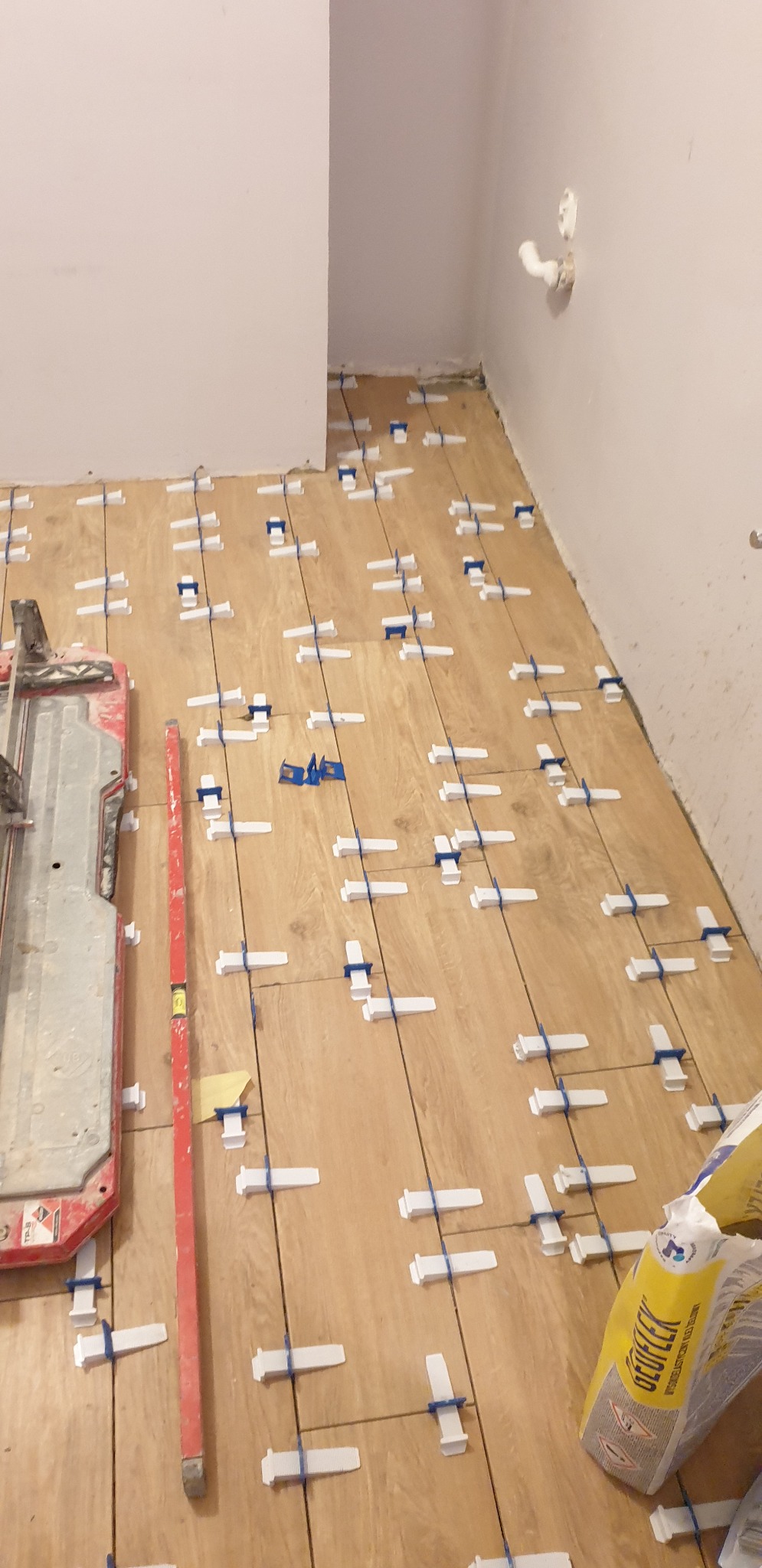 Podłoga w trakcie układania paneli imitujących drewno, z użyciem białych i niebieskich klinów dystansowych; widoczna poziomica, paczka kleju i narzędzie do cięcia paneli.
