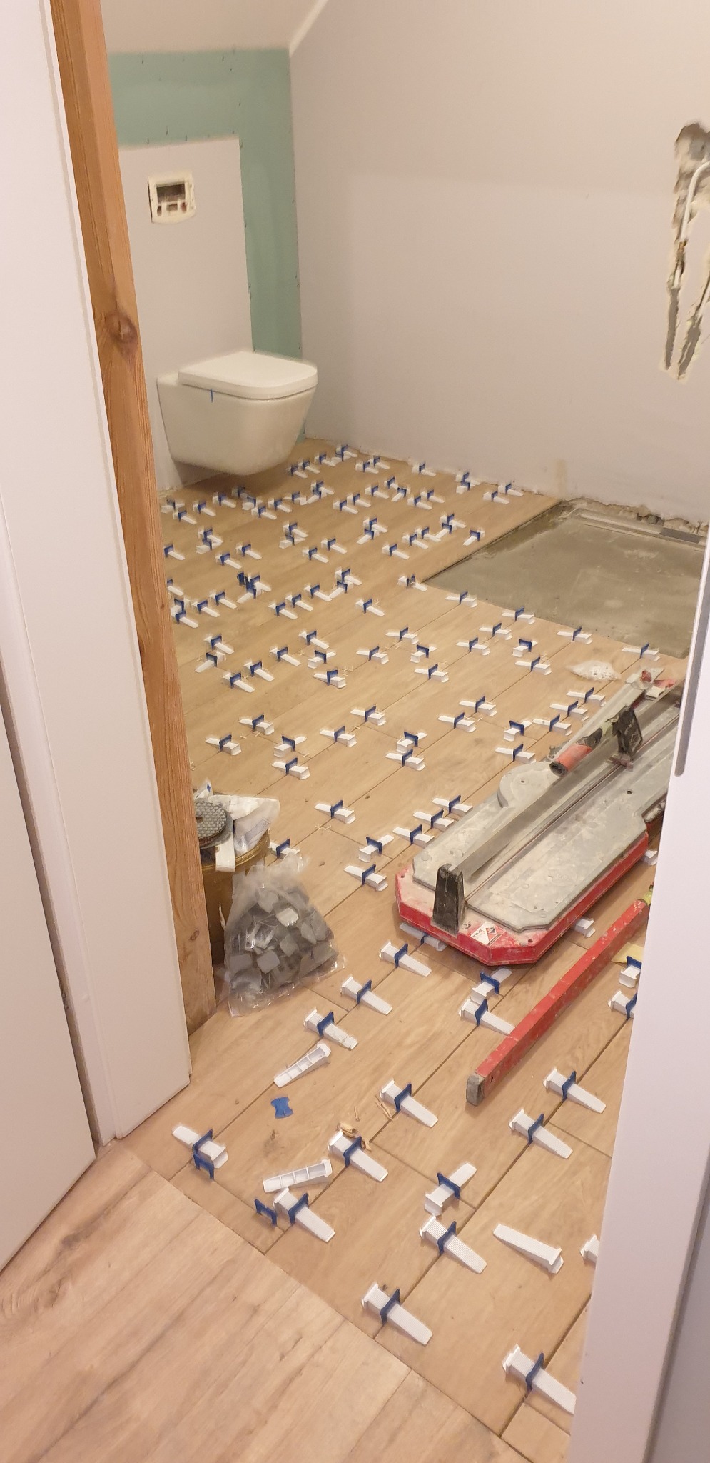 Układanie płytek podłogowych w łazience, widoczne krzyżyki dystansowe, poziomica i przecinarka do płytek, z zamontowaną toaletą podwieszaną.