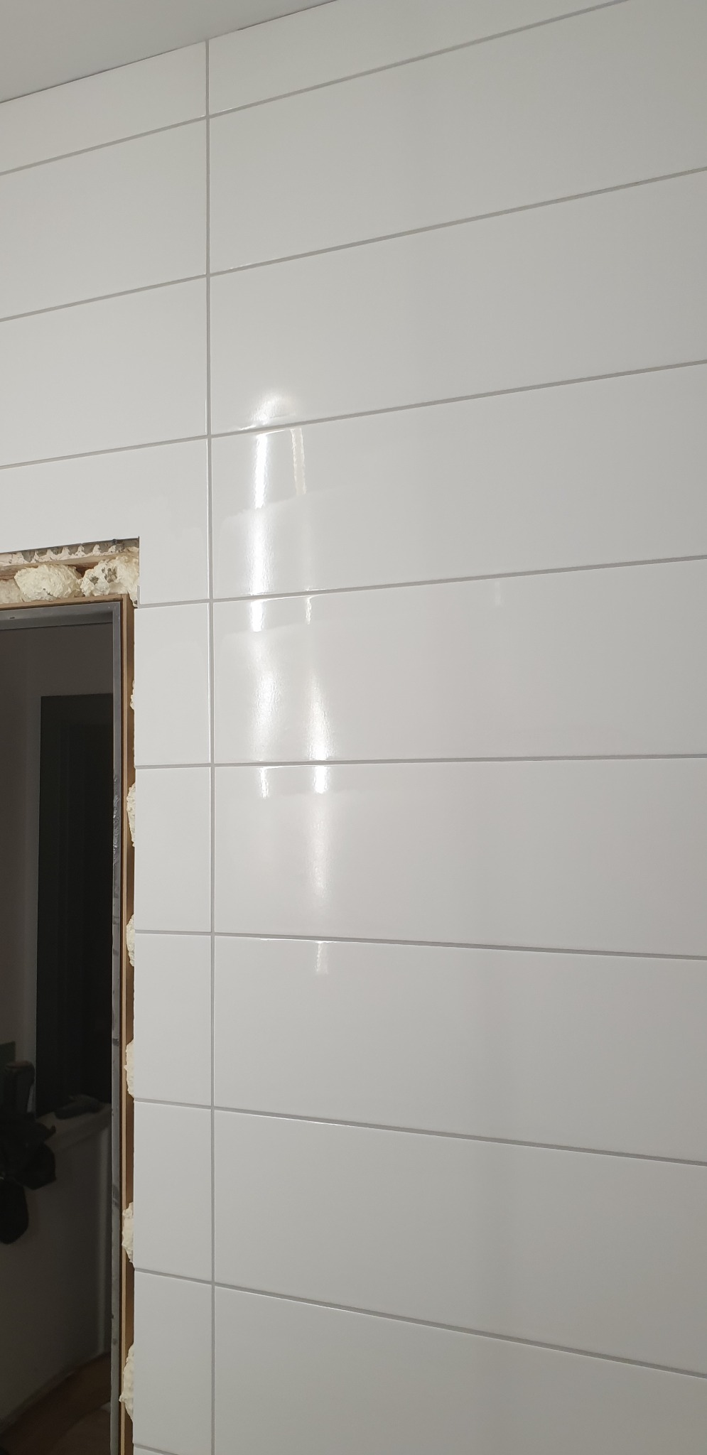 Ściana wyłożona błyszczącymi, białymi, poziomymi płytkami ceramicznymi z widocznym fragmentem ościeżnicy drzwiowej wypełnionej pianką montażową.