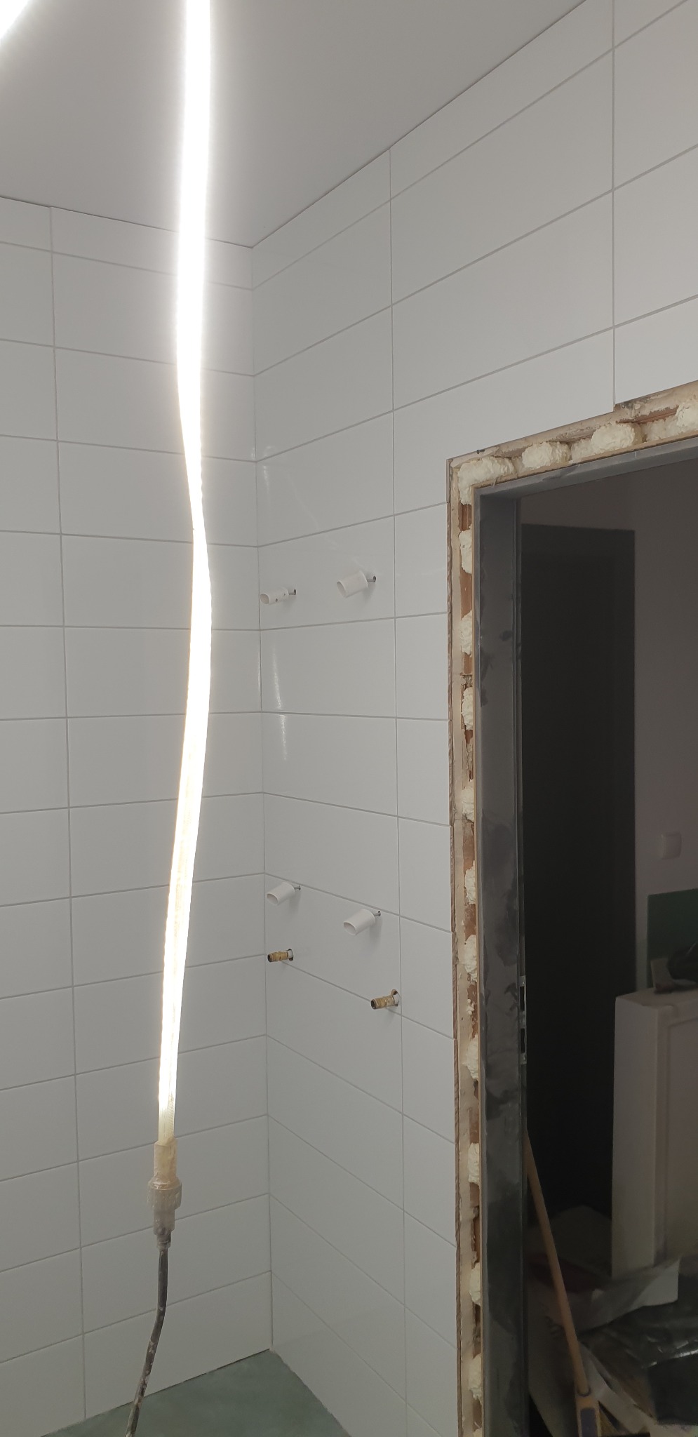 Białe, kwadratowe płytki w łazience w trakcie montażu, widoczne punkty instalacji wodnej i fragment ościeżnicy zabezpieczonej pianką montażową, oświetlenie robocze