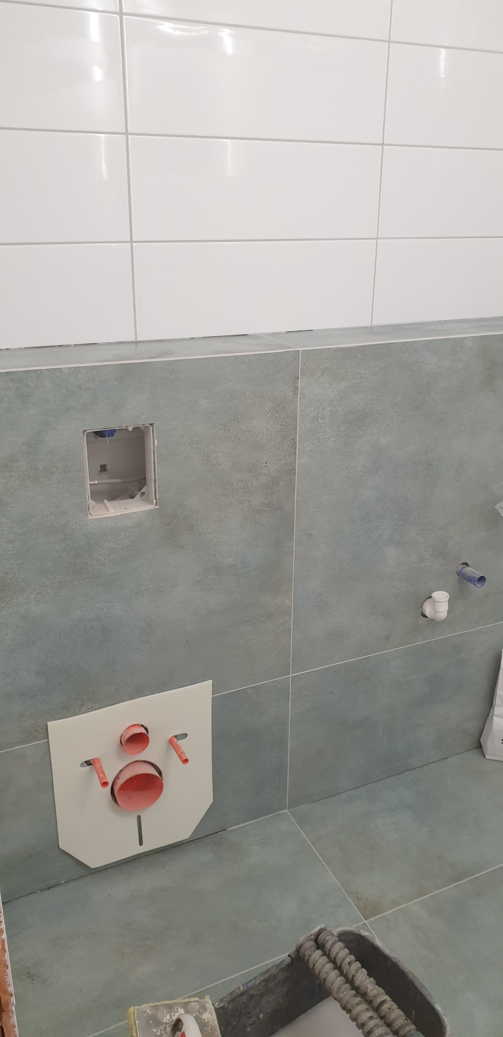 Ściana łazienki z szarymi płytkami imitującymi kamień, częściowo wykończona białymi płytkami, z widoczną instalacją podtynkową WC i otwartym otworem na przyłącza. W tle widoczne elementy instalacji...