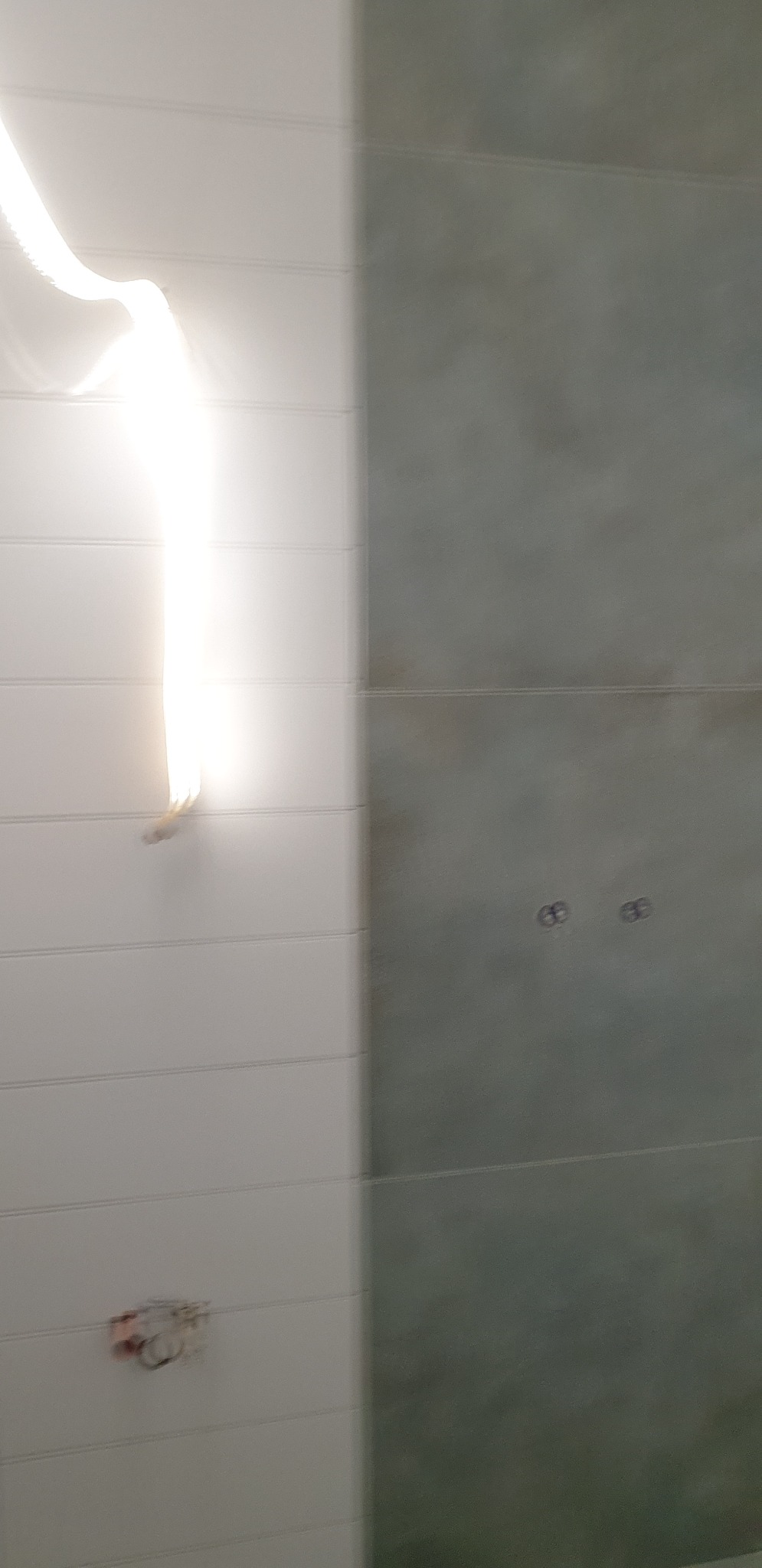 Ściana w łazience wyłożona dwoma rodzajami płytek: białe, poziome panele oraz szare, kwadratowe płytki. Widoczne otwory po instalacji oraz odbicie światła.