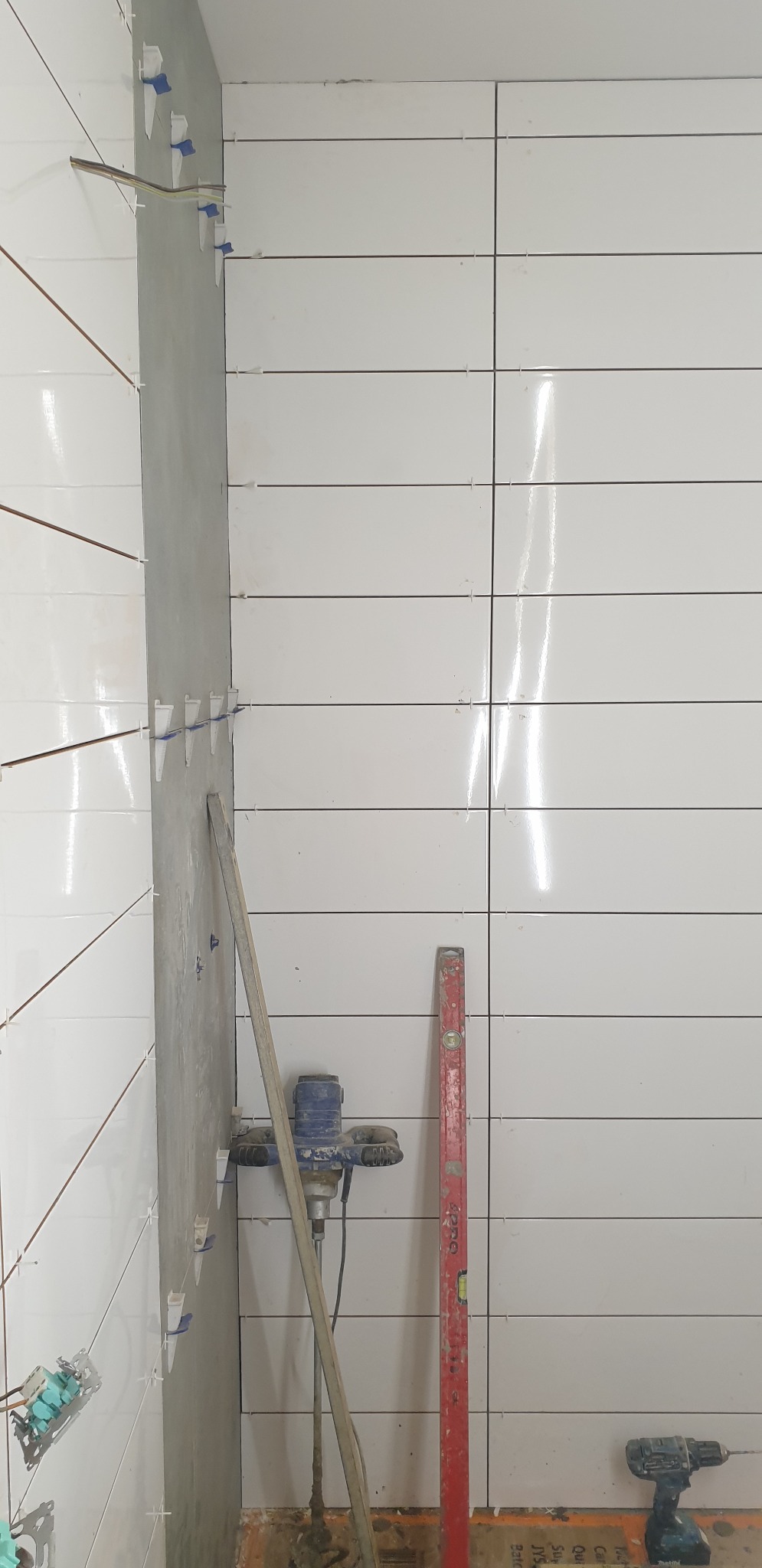 Świeżo ułożone, białe, poziome płytki w łazience, z widocznymi krzyżykami dystansowymi, poziomica, mieszadło do zaprawy i wiertarka na podłodze.