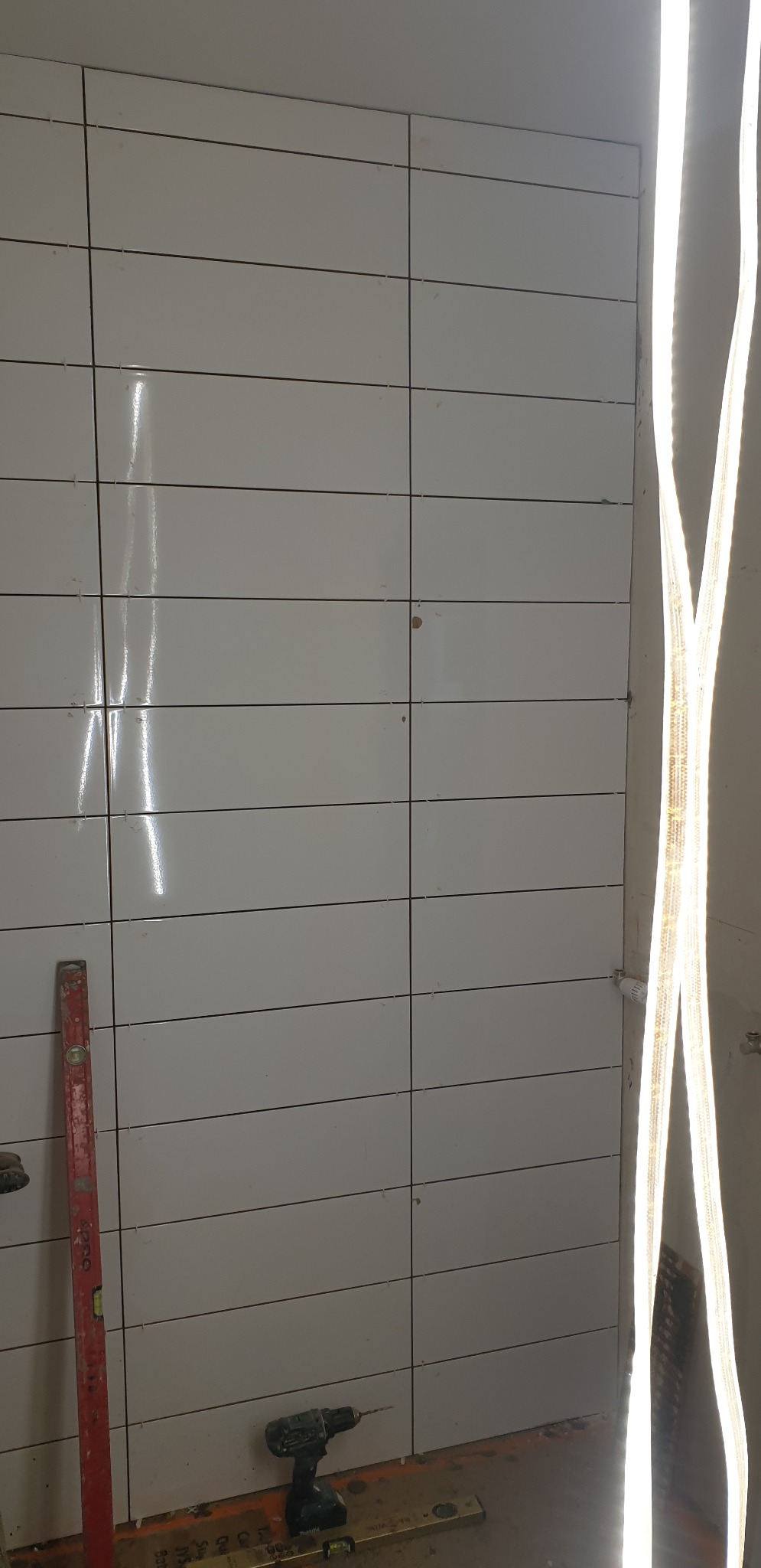 Ściana wyłożona białymi, poziomymi płytkami ceramicznymi w trakcie montażu, widoczny poziomica, wiertarka akumulatorowa i szczelina dylatacyjna przy podłodze.
