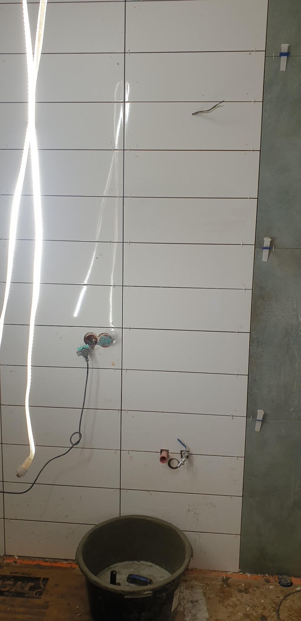 Ściana łazienki w trakcie układania białych, poziomych płytek z widocznymi krzyżykami dystansowymi, niepodłączonymi kranami i wiaderkiem z zaprawą na podłodze. W tle fragment ściany z szarą płytką.