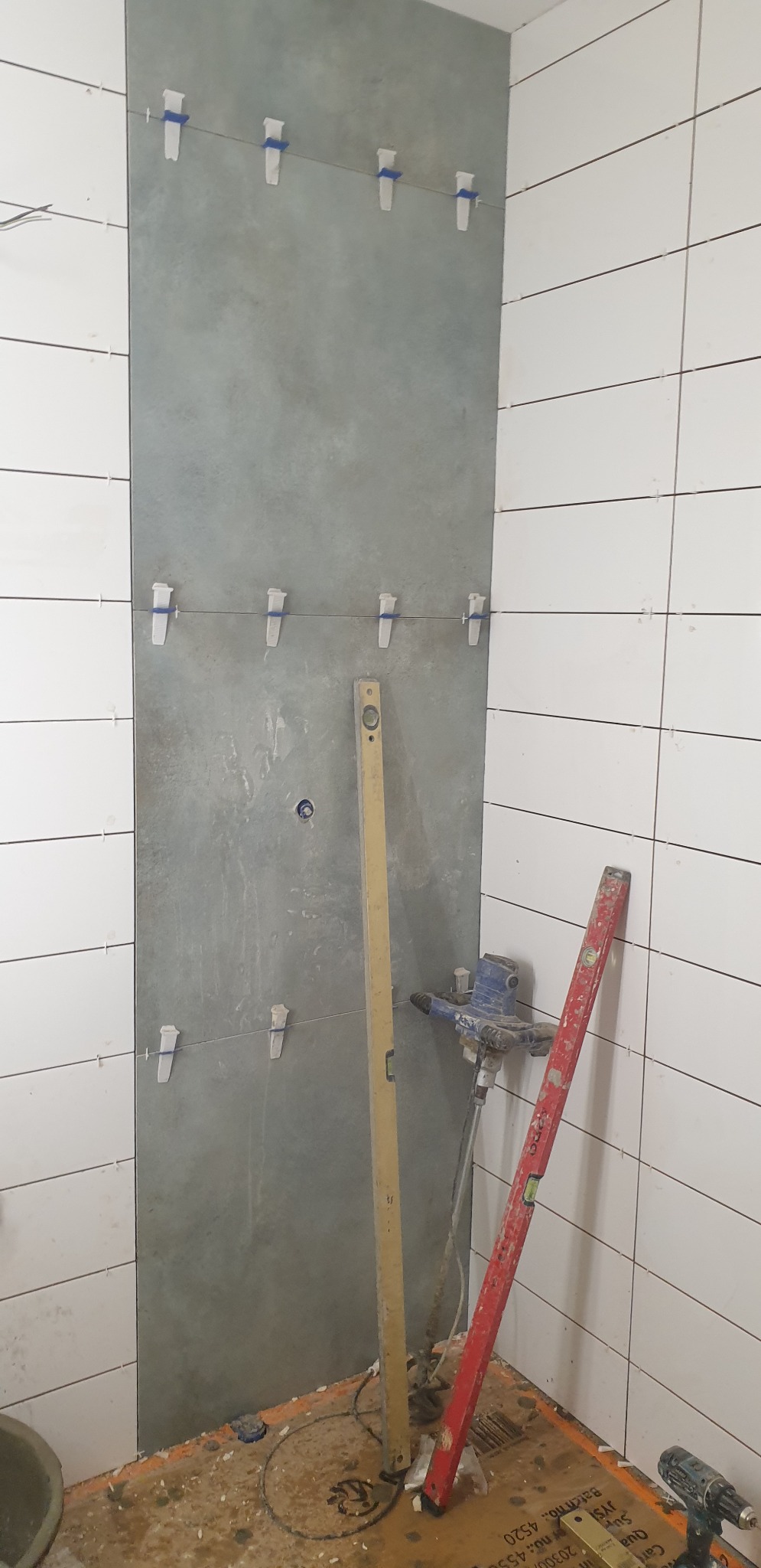 Układanie płytek w łazience: szare płytki na ścianie mocowane za pomocą poziomicy i krzyżyków dystansowych, na podłodze widoczne narzędzia i resztki kleju.