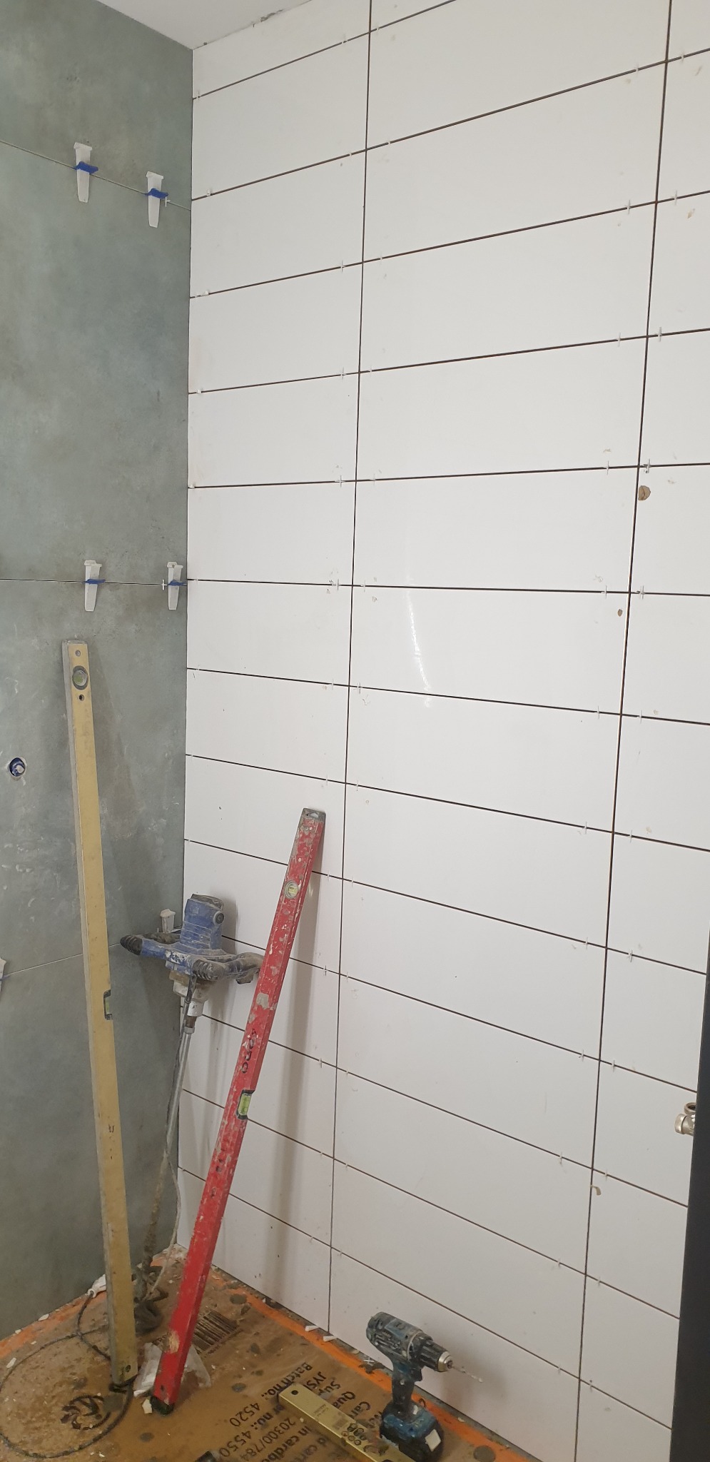 Białe, prostokątne płytki ułożone poziomo na ścianie łazienki, z widocznymi krzyżykami dystansowymi; obok ściany z surowym betonem, poziomice, mieszadło do zaprawy i wkrętarka na podłodze.