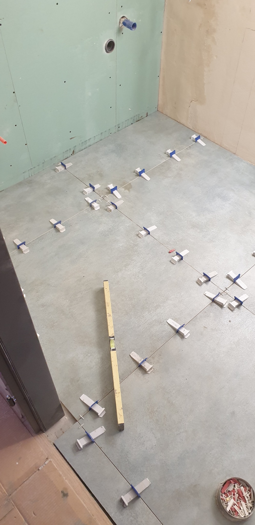 Podłoga w trakcie układania płytek, widoczne krzyżyki dystansowe i poziomica, ściany przygotowane pod wykończenie w łazience.