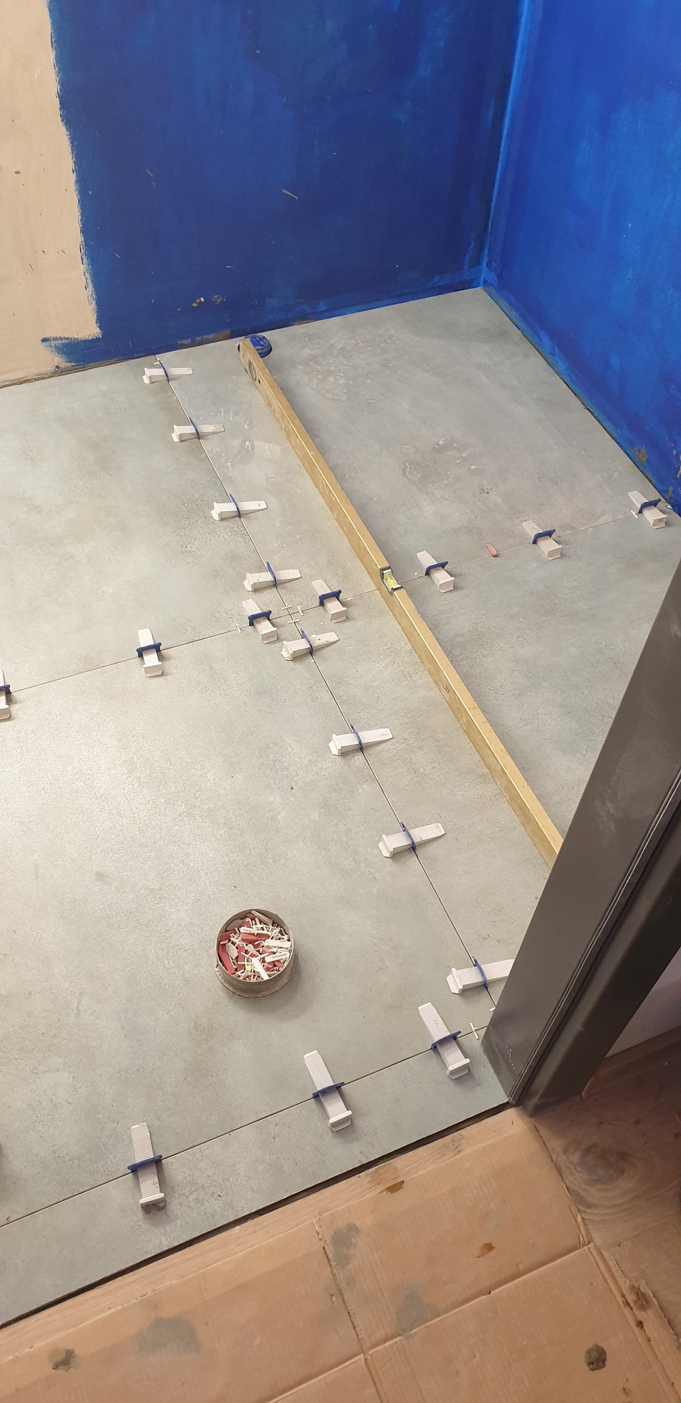 Podłoga w trakcie układania płytek, z użyciem poziomicy i klipsów dystansowych, widoczny pojemnik z klinami, ściany częściowo pomalowane na niebiesko.