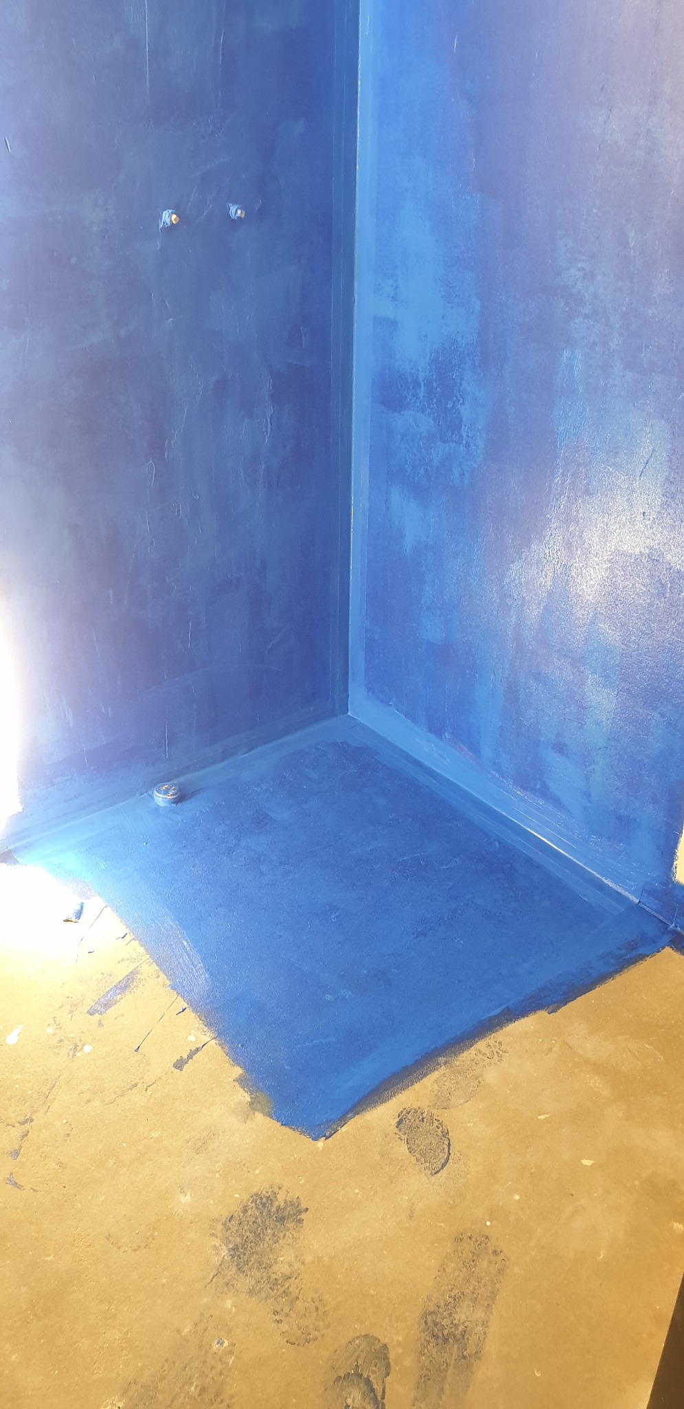 Niebieska hydroizolacja w rogu łazienki, widoczne wystające rury instalacji wodnej i odpływ podłogowy, podłoga przed wykończeniem.