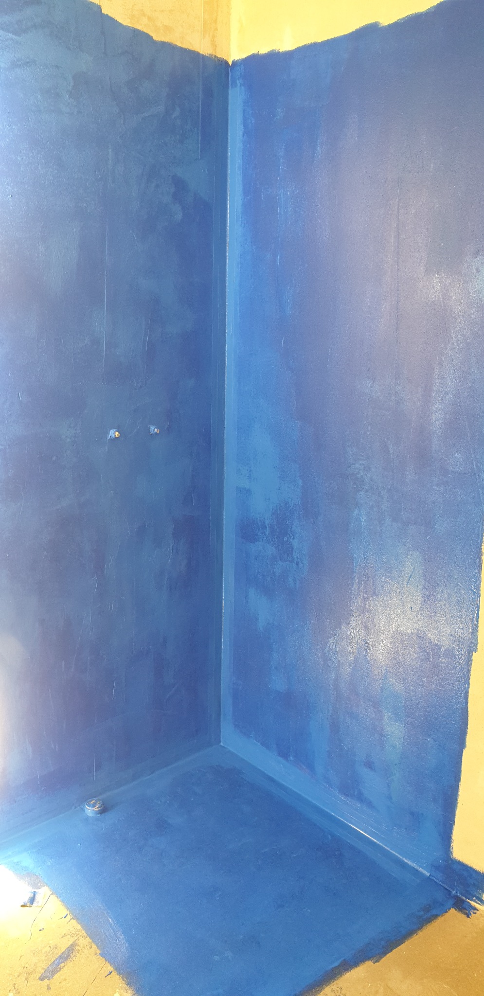 Niebieska hydroizolacja w rogu prysznica, widoczne rury i odpływ podłogowy, przygotowanie do dalszych prac wykończeniowych.