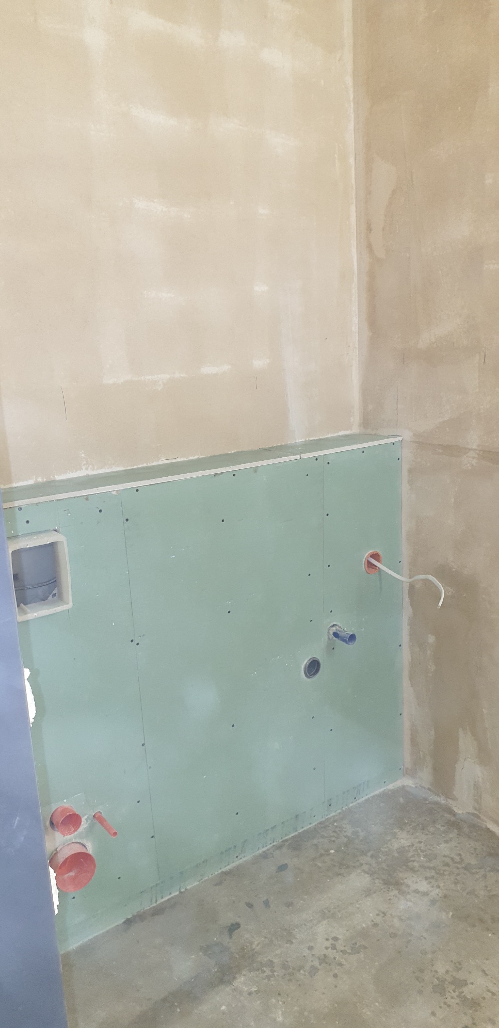 Surowa łazienka w trakcie przygotowania do wykończenia: zielone płyty gipsowo-kartonowe zamontowane na ścianach, widoczne instalacje wodne i elektryczne, surowe ściany w odcieniach beżu, betonowa...