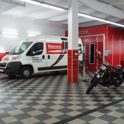 Szewczyk Firma Motoryzacyjna - Warsztat tapicerski z białą furgonetką z logo firmy Szewczyk i motocyklem na szachownicowej podłodze, na tle czerwonej kabiny lakierniczej.
