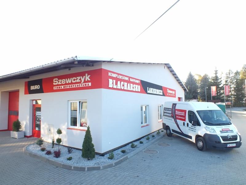 Budynek firmy motoryzacyjnej z napisem 'Szewczyk' i zaparkowany biały van z logo firmy.