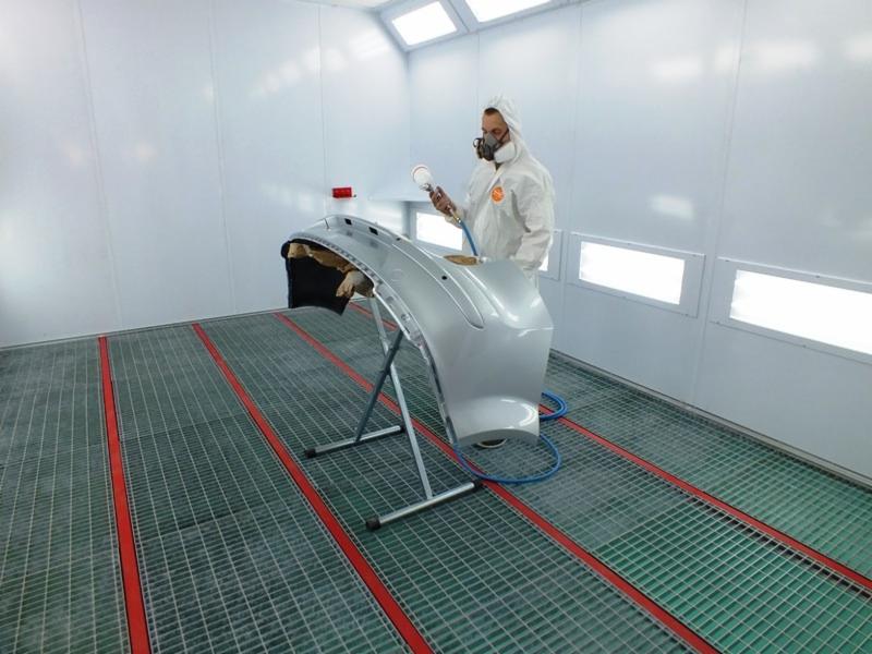 Lakiernik w kombinezonie ochronnym maluje srebrny zderzak samochodowy w kabinie lakierniczej, widoczne kraty podłogowe i oświetlenie sufitowe.