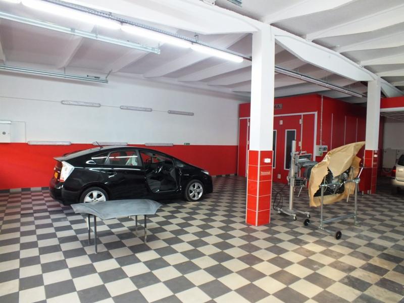 Czarne auto z otwartymi drzwiami w warsztacie z podłogą w szachownicę, widoczna maska na stojaku i kabina lakiernicza w tle.