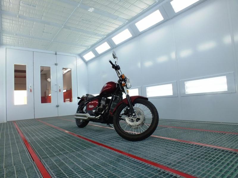 Bordowy motocykl w kabinie lakierniczej z kratownicą podłogową i oświetleniem sufitowym.