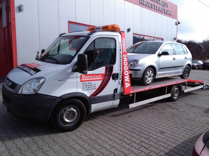 Laweta z srebrnym samochodem osobowym na platformie, zaparkowana przed budynkiem z napisem REJESTRACJE, widoczne logo firmy Szewczyk Pomoc Drogowa.